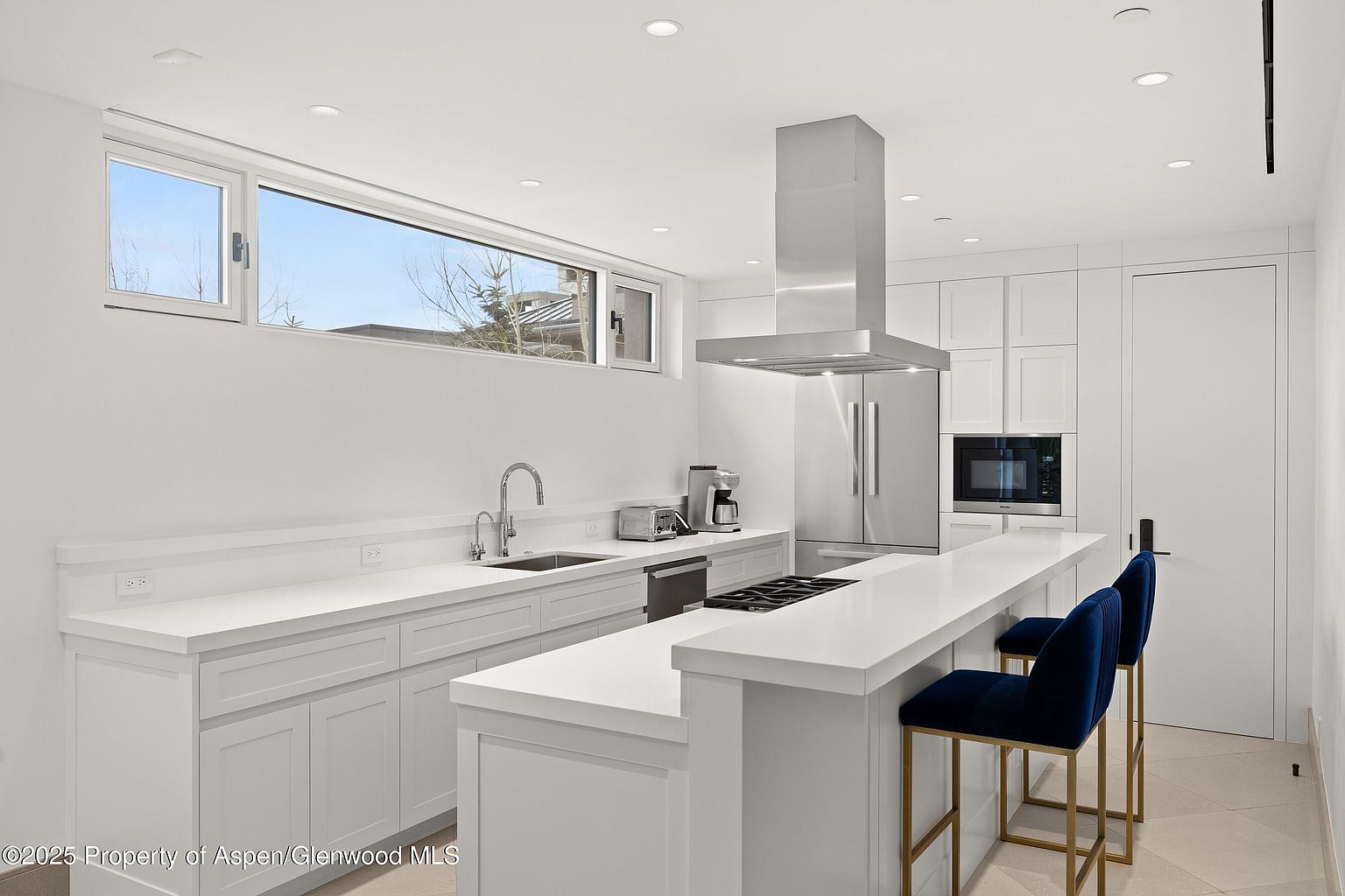 This is a bright, modern kitchen with white cabinetry and countertops. Stainless steel appliances, including a refrigerator and range hood, add a sleek touch. Two blue velvet bar stools are positioned at the island, offering seating, and a large window provides natural light.