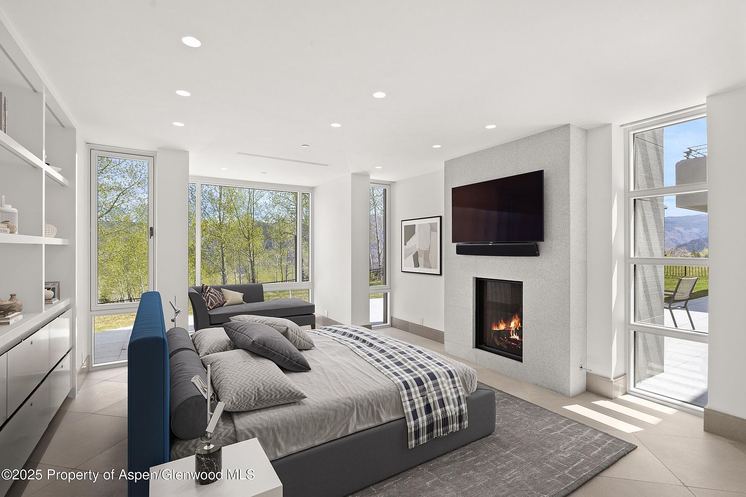 This is a spacious primary bedroom featuring a modern design with a fireplace and a large television mounted above it. The room has large windows offering views of the surrounding landscape, and a seating area is present. The color palette is neutral with accents of blue, creating a serene and luxurious atmosphere.