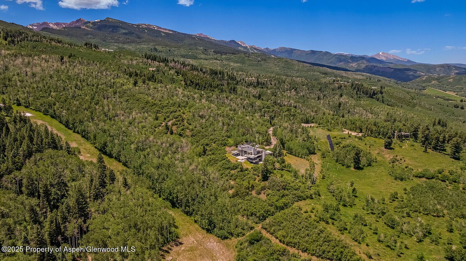 This aerial view showcases a contemporary home nestled amidst a lush, green landscape with mountains in the background. The house features a modern architectural design with multiple levels and a flat roof. The surrounding area is heavily wooded, providing privacy and a sense of seclusion, making it an ideal retreat.