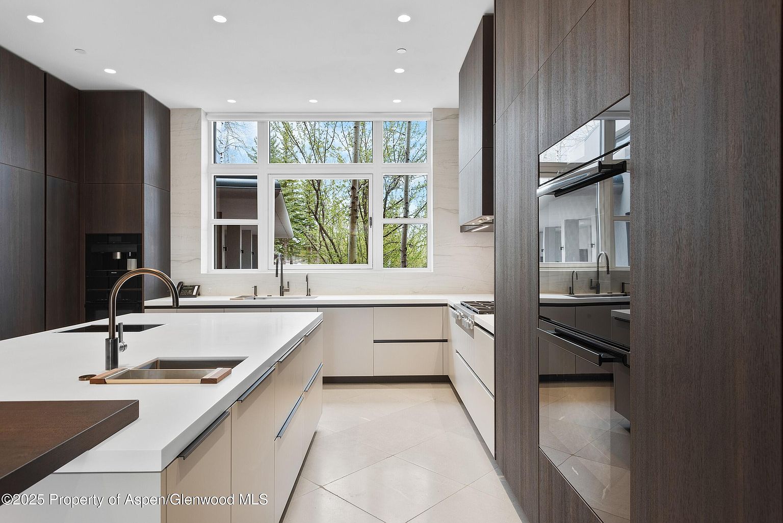This is a modern kitchen with dark wood and light beige cabinetry, featuring a large island with a white countertop and a stainless steel sink. A large window provides natural light and a view of the outdoors. The kitchen is equipped with modern appliances, including a built-in oven and a cooktop.