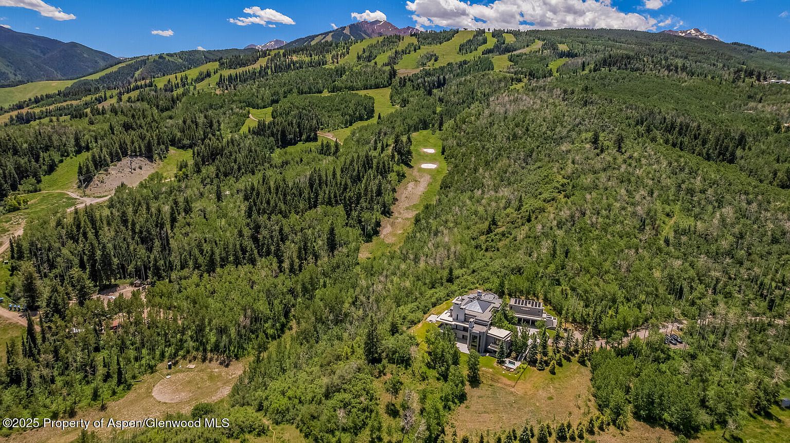 This aerial view showcases a luxurious modern home nestled amidst a lush, green landscape with a mountain backdrop. The architecture features clean lines and a sophisticated design, blending seamlessly with the natural surroundings. The property includes manicured grounds and appears to offer privacy and seclusion.