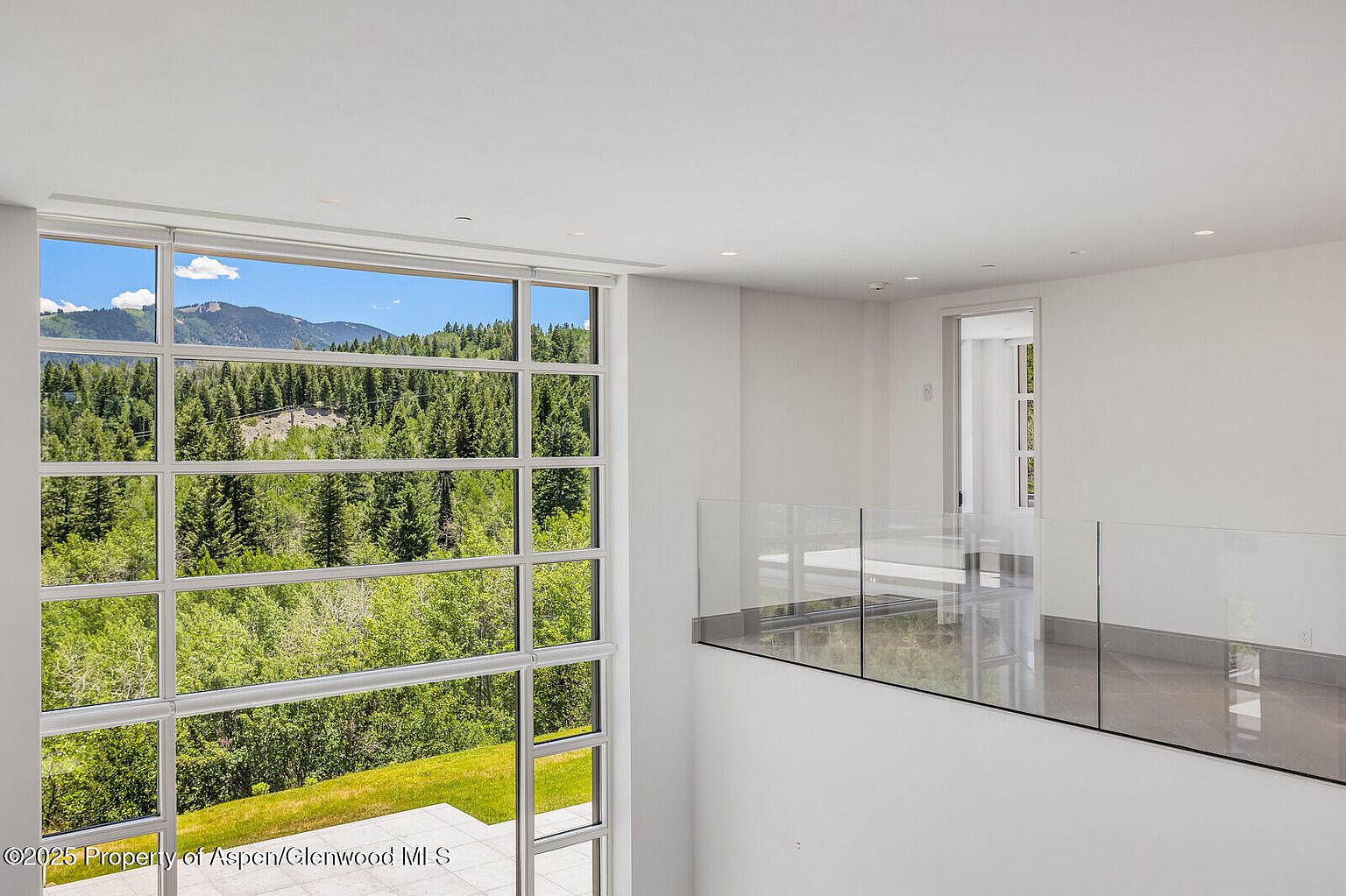 This interior shot showcases a modern hallway or landing area with a large window offering a scenic view of mountains and trees. The space features clean white walls, a glass railing, and a glimpse of another room through a doorway. The design emphasizes natural light and a minimalist aesthetic.