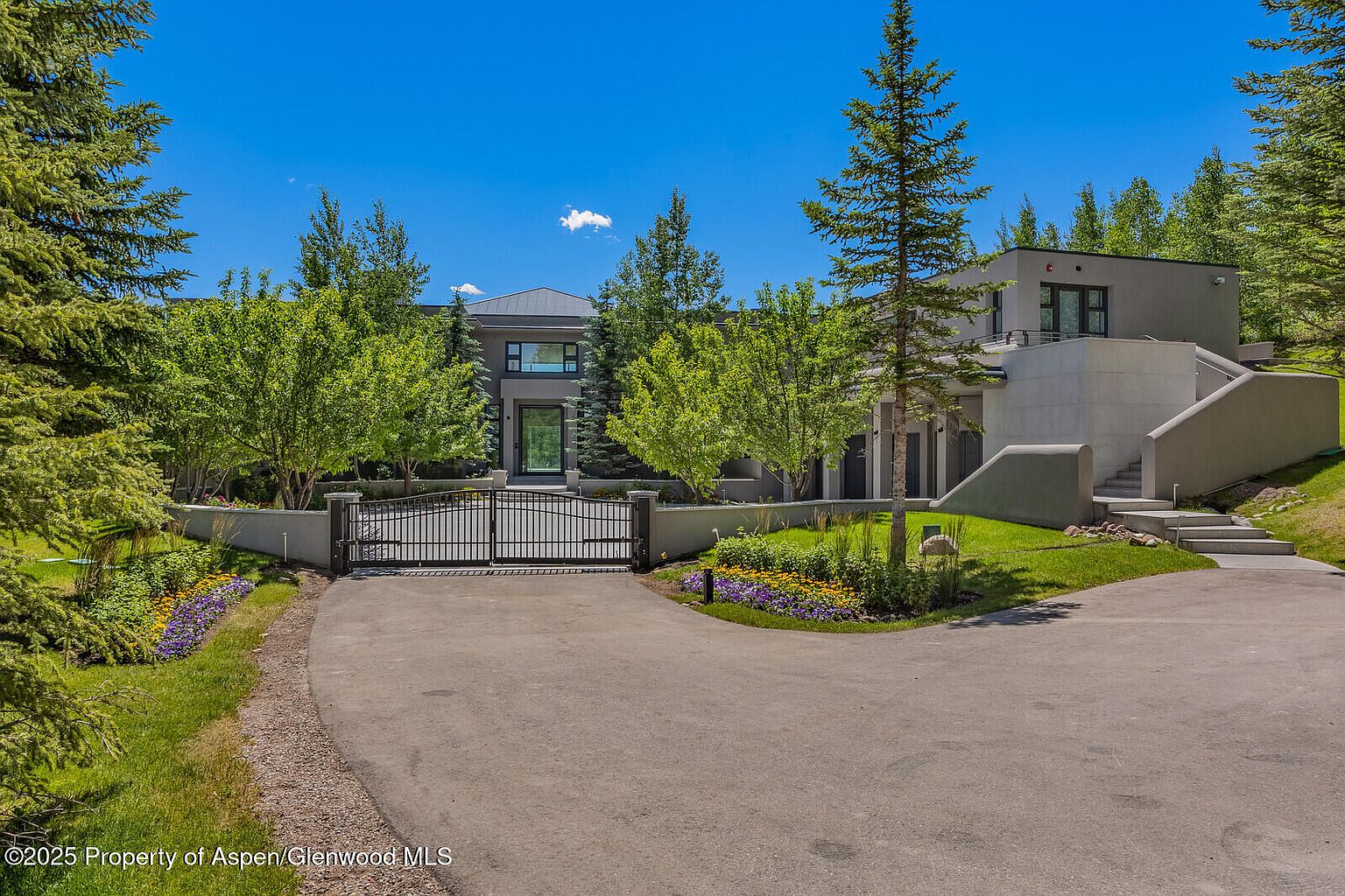 This is a front exterior view of a modern home with a gated entrance. The property features a combination of architectural styles, including a flat-roofed section and a gabled section. Landscaping includes mature trees, flower beds, and a well-maintained lawn, enhancing the property's curb appeal.