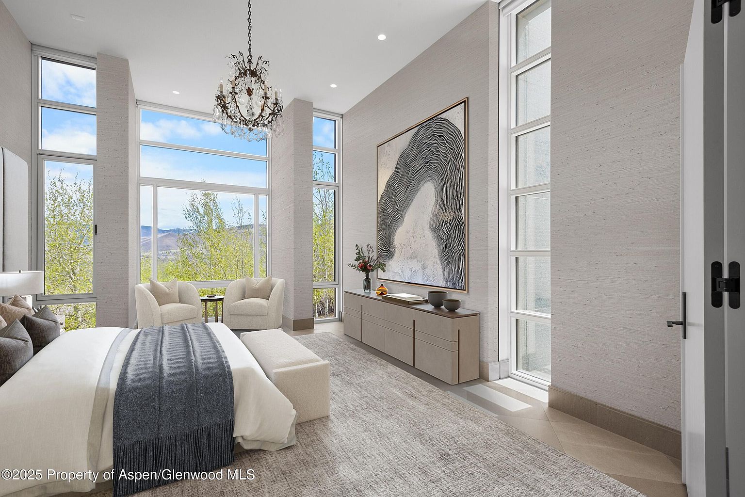 This is a luxurious primary bedroom featuring a large bed with a gray throw blanket, two armchairs facing a window with a mountain view, and a console table with artwork above it. The room is well-lit with natural light from the large windows and features a chandelier, creating an elegant and inviting atmosphere. The neutral color palette and textured walls add to the room's sophisticated style.