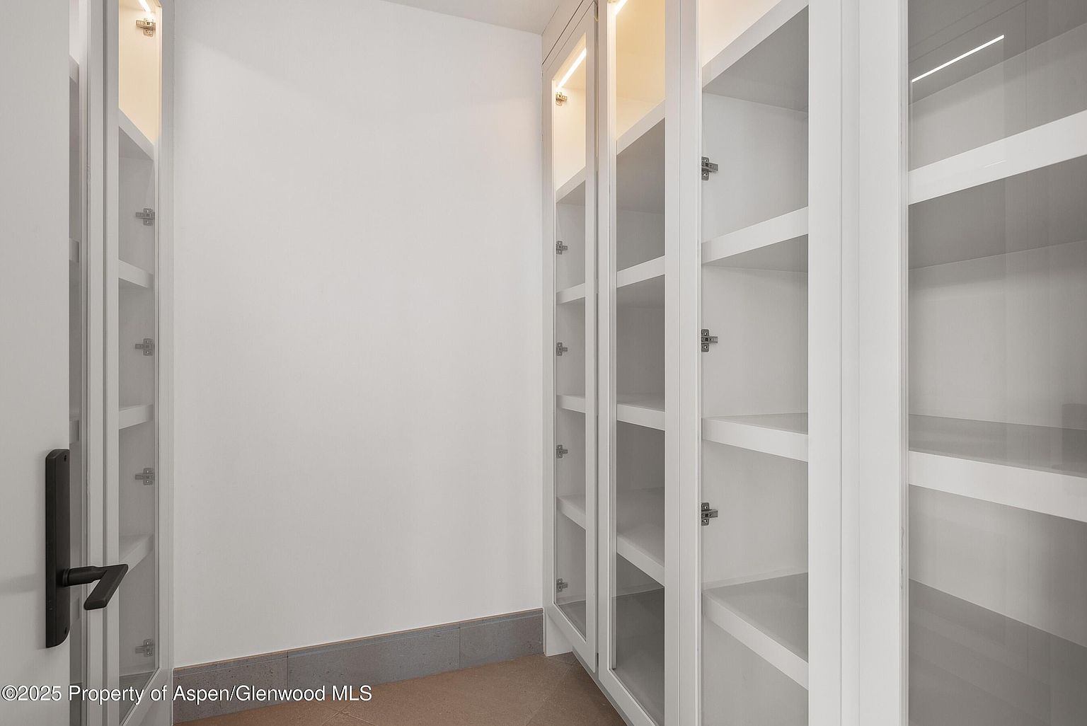 This image showcases a well-organized walk-in closet with white shelving and glass doors, illuminated by subtle lighting. The closet features multiple shelves for storage and a clean, minimalist design. The overall impression is one of luxury and efficient use of space, perfect for showcasing in a real estate listing.