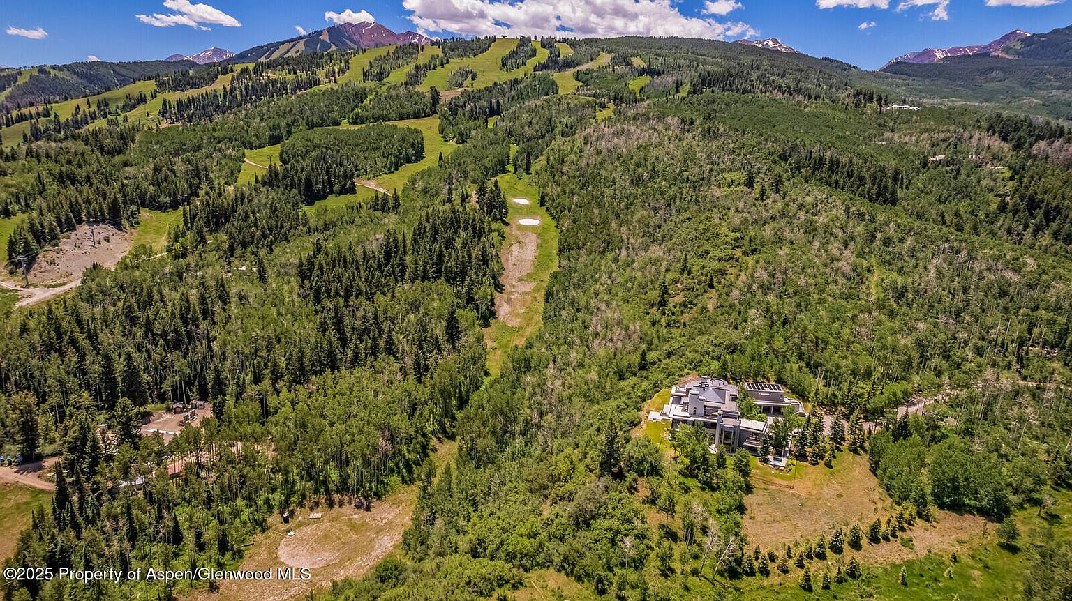 This aerial view showcases a luxurious estate nestled amidst lush greenery and rolling hills. The property features a large, modern home with multiple wings and a meticulously landscaped yard. The surrounding landscape includes dense forests and open ski runs, creating a sense of privacy and natural beauty.