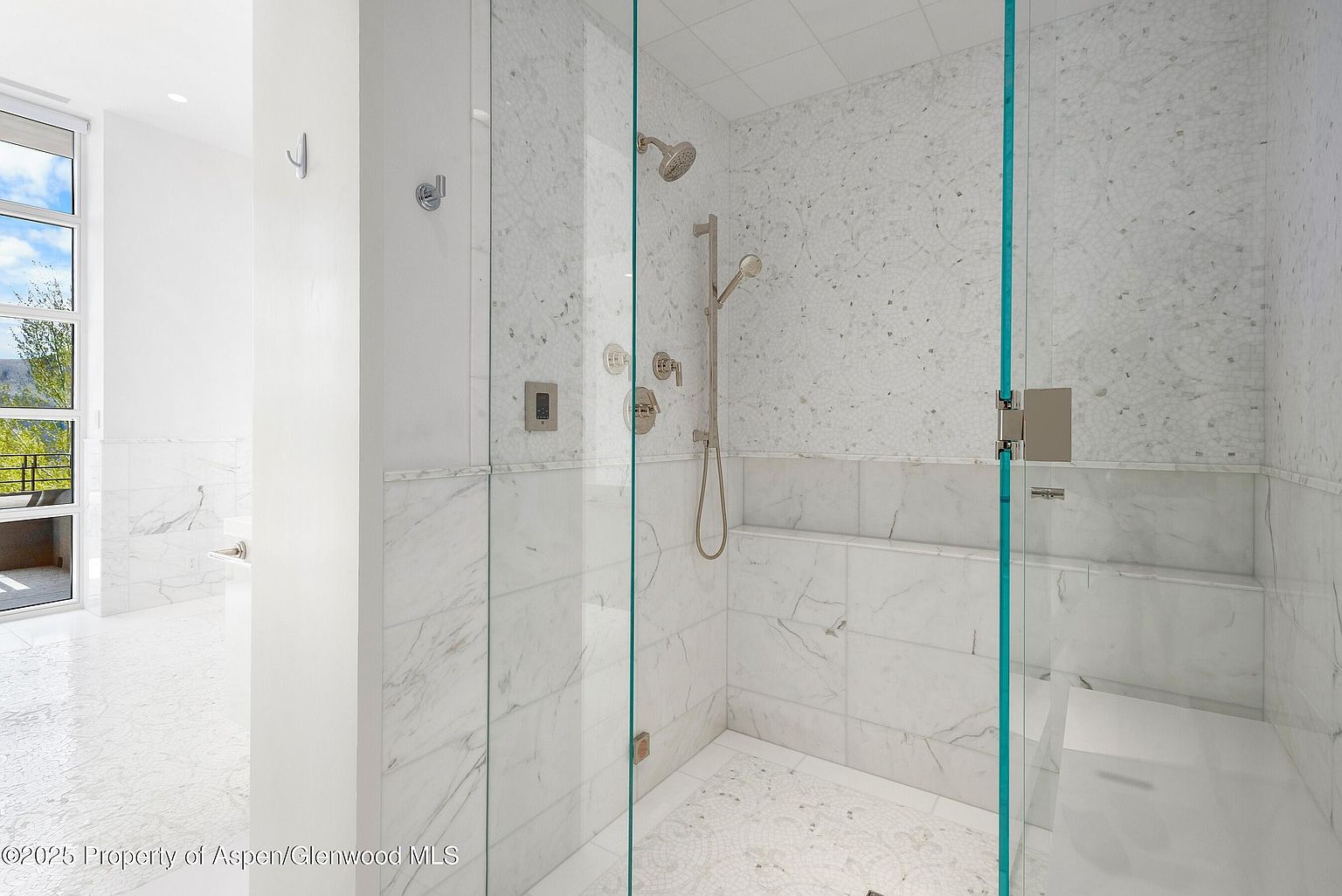 This is a bright and luxurious primary bathroom featuring a glass-enclosed shower with marble walls and mosaic accents. The shower includes multiple showerheads and sleek fixtures. The bathroom has a clean, modern aesthetic with a focus on high-end materials and finishes, creating a spa-like atmosphere.