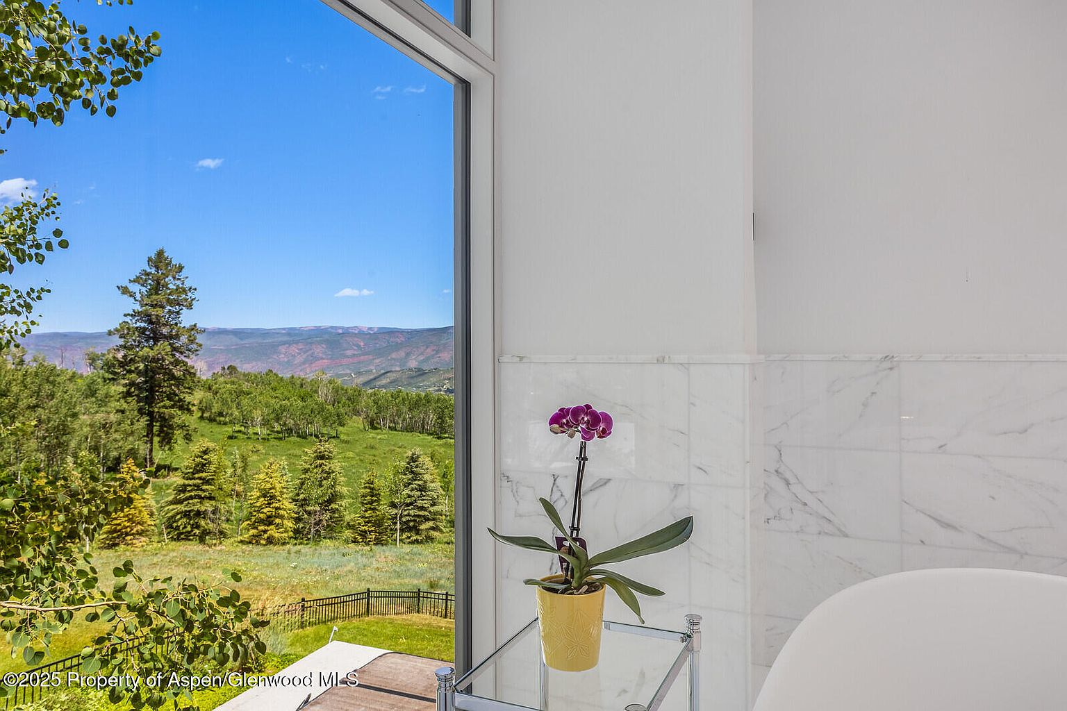 This image showcases a bright primary bathroom with marble tiling and a view of a lush, green landscape through a large window. A purple orchid in a yellow pot sits on a glass table, adding a pop of color to the otherwise neutral palette. The overall impression is one of luxury and tranquility, perfect for relaxation.