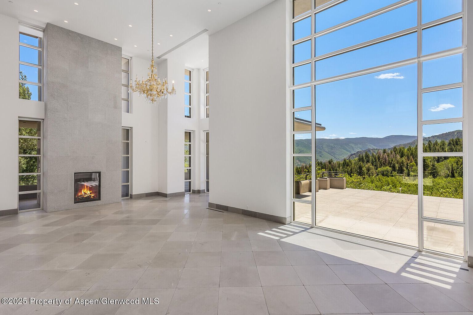 This is a spacious living room featuring a modern design with high ceilings and large windows offering scenic mountain views. The room includes a fireplace with a stone surround and a decorative chandelier. The neutral color palette and tile flooring create a bright and airy atmosphere, perfect for entertaining or relaxing.