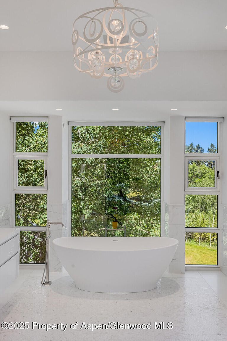 This is a bright and airy primary bathroom featuring a freestanding white bathtub positioned in front of a large window offering a view of lush greenery. The room is decorated in a minimalist style with white walls, white tile flooring, and a modern chandelier, creating a serene and spa-like atmosphere. The perspective is from the doorway, showcasing the tub as the focal point.