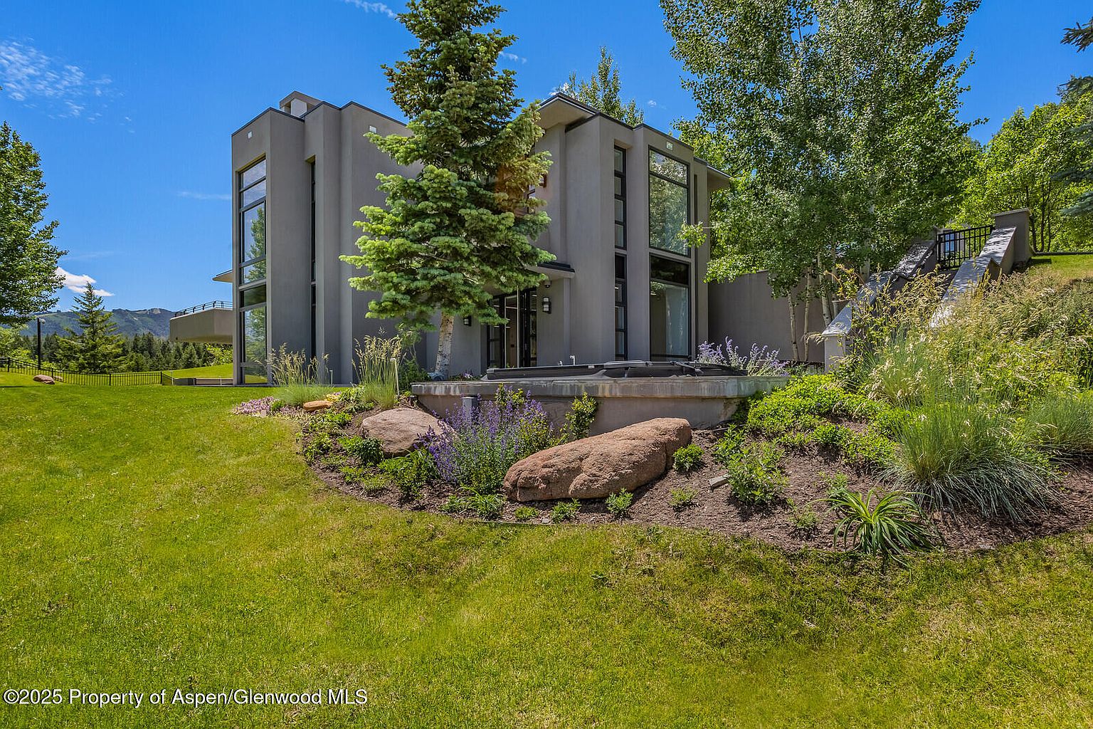 This image showcases the exterior of a modern home with a well-manicured yard and garden. The architecture features clean lines and large windows, complemented by mature trees and landscaping. A stone-lined garden bed with various plants and large rocks adds visual interest to the property.