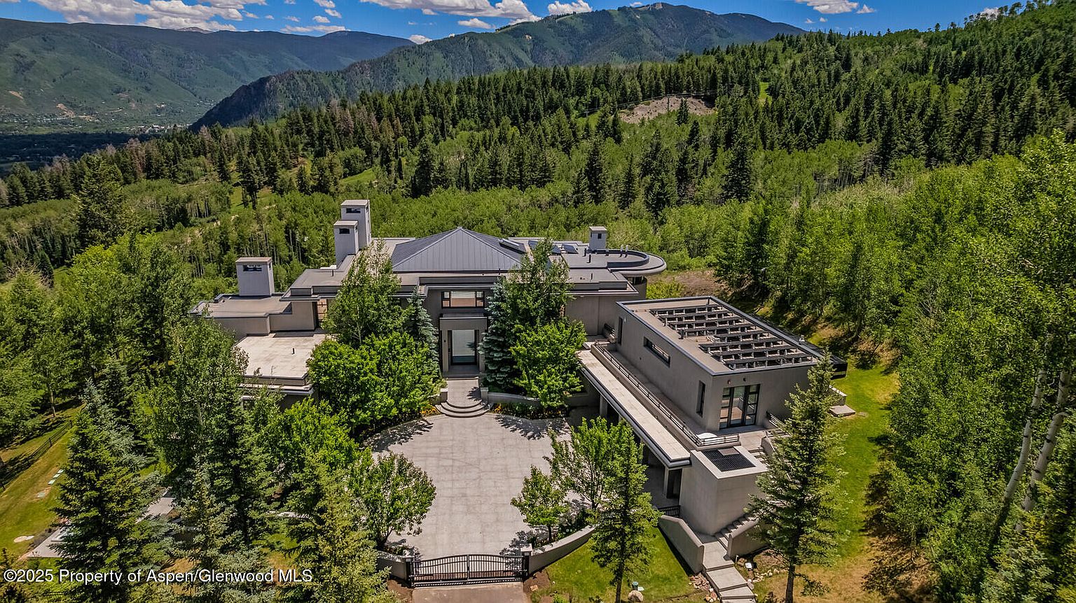 This aerial view showcases a luxurious modern home nestled in a lush, mountainous landscape. The property features a contemporary architectural design with clean lines, a flat roof, and a spacious driveway. The surrounding mature trees provide privacy and enhance the natural beauty of the setting, creating an exclusive and serene atmosphere.