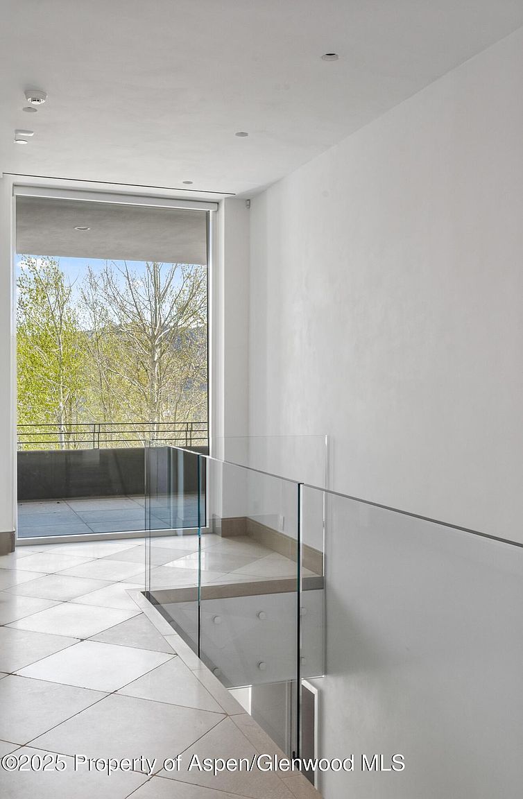 This interior shot showcases a modern hallway and staircase with a glass railing, emphasizing clean lines and a minimalist aesthetic. The flooring features large, light-colored tiles arranged in a diamond pattern, while a large window provides natural light and a view of the outdoors. The overall impression is bright, airy, and sophisticated, highlighting the contemporary design of the home.