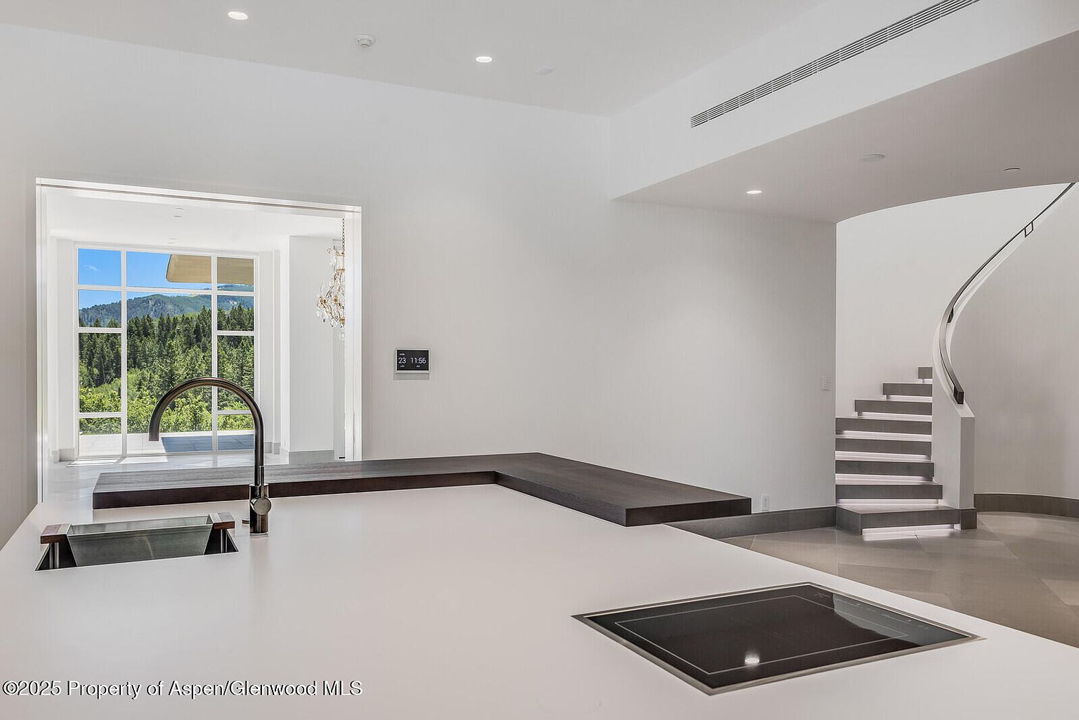This is a modern kitchen with a minimalist design, featuring a white countertop with a built-in sink and cooktop. A dark wood extension serves as a bar or serving area, and a large window offers a view of the surrounding landscape. A modern staircase with integrated lighting is visible in the background, adding to the contemporary aesthetic.