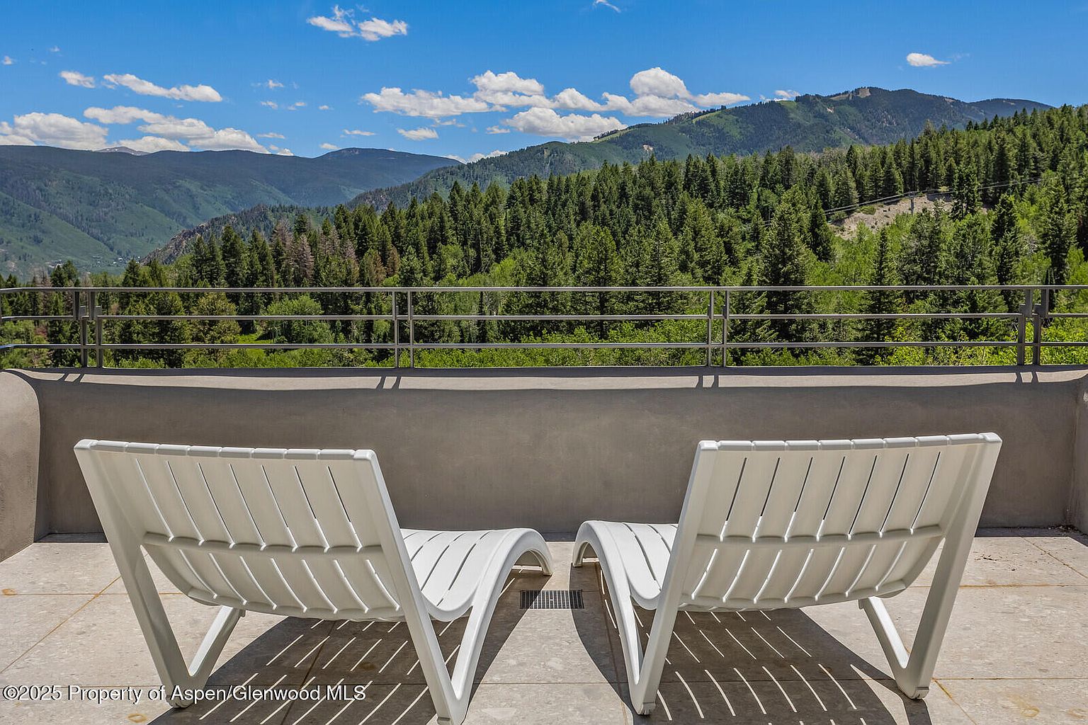 This image showcases a serene balcony or patio setting with two white lounge chairs positioned to take in the breathtaking mountain views. The balcony features a neutral-toned concrete wall and a metal railing, providing a modern yet understated aesthetic. The lush greenery of the surrounding trees and the clear blue sky with scattered clouds create a tranquil and inviting atmosphere, perfect for relaxation and enjoying the natural landscape.