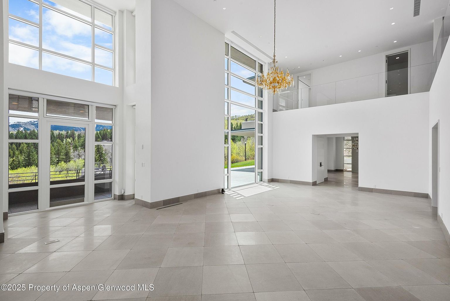 This is a spacious and modern living room featuring high ceilings, large windows offering natural light, and a neutral color palette. The room has tile flooring and a contemporary chandelier, creating an open and airy atmosphere. A second-floor balcony overlooks the living area, adding architectural interest.