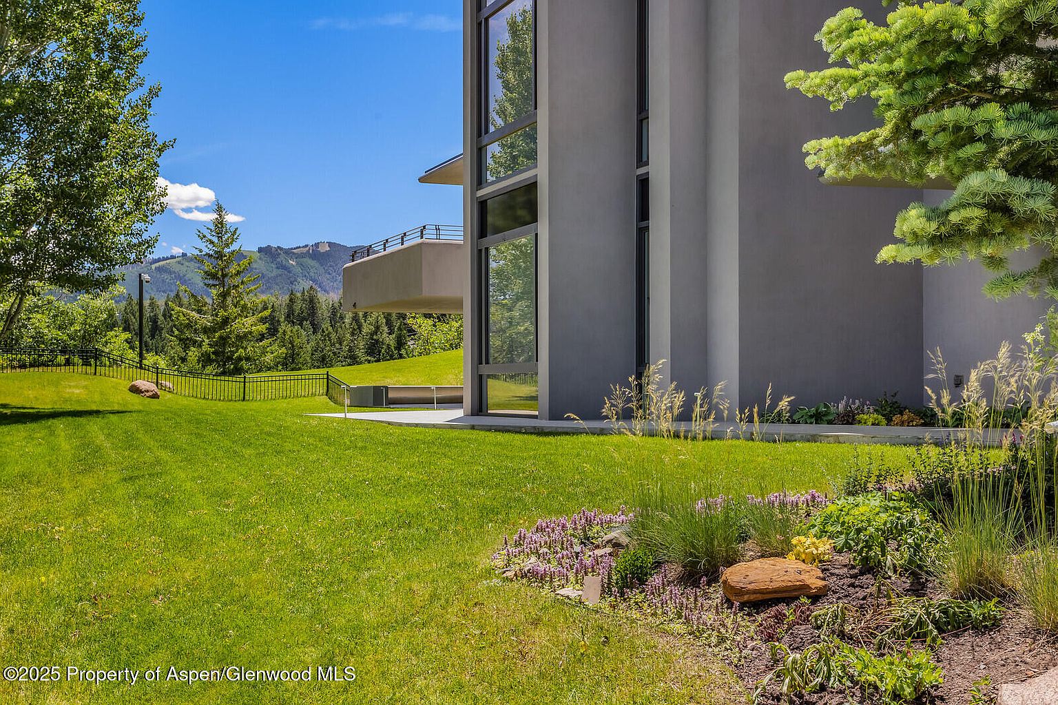 This image showcases the exterior of a modern home, focusing on the well-maintained yard and garden. The building features large windows and a balcony, blending seamlessly with the natural landscape. Lush green grass, trees, and a flower bed with rocks add to the property's appeal, creating a serene and inviting atmosphere.