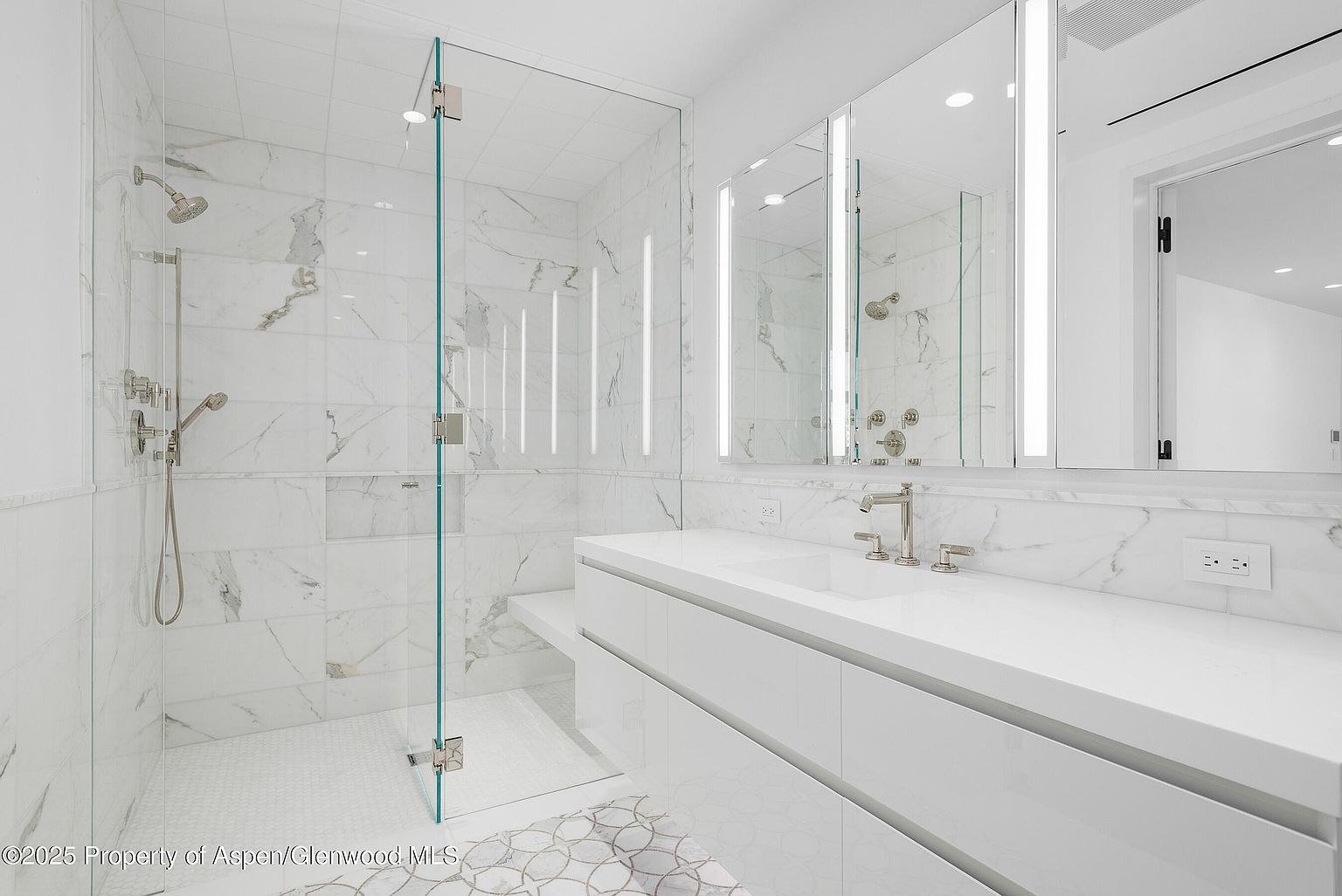 This is a bright and modern primary bathroom featuring a glass-enclosed shower with marble tile walls and a mosaic tile floor. The vanity has a white countertop and sleek white cabinets, complemented by a large mirror with integrated lighting. The overall impression is clean, luxurious, and well-lit.