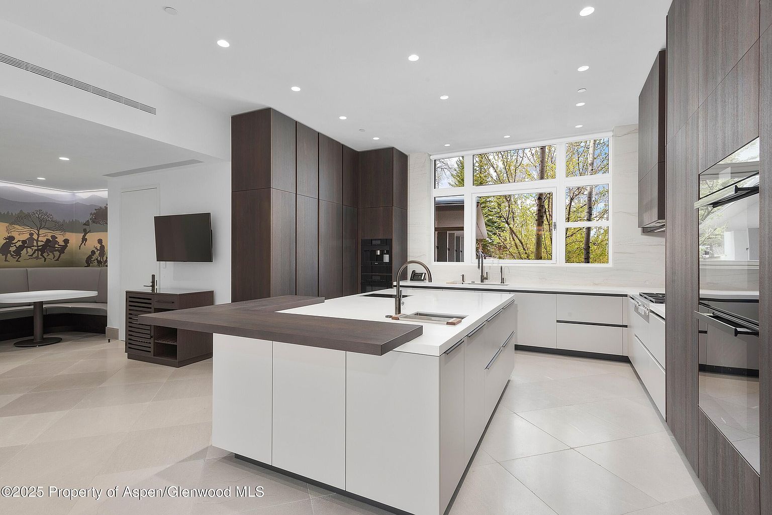 This is a modern kitchen with a large island featuring a white countertop and white cabinets. The kitchen also has dark wood cabinets and stainless steel appliances. A large window provides natural light and a view of the outdoors.