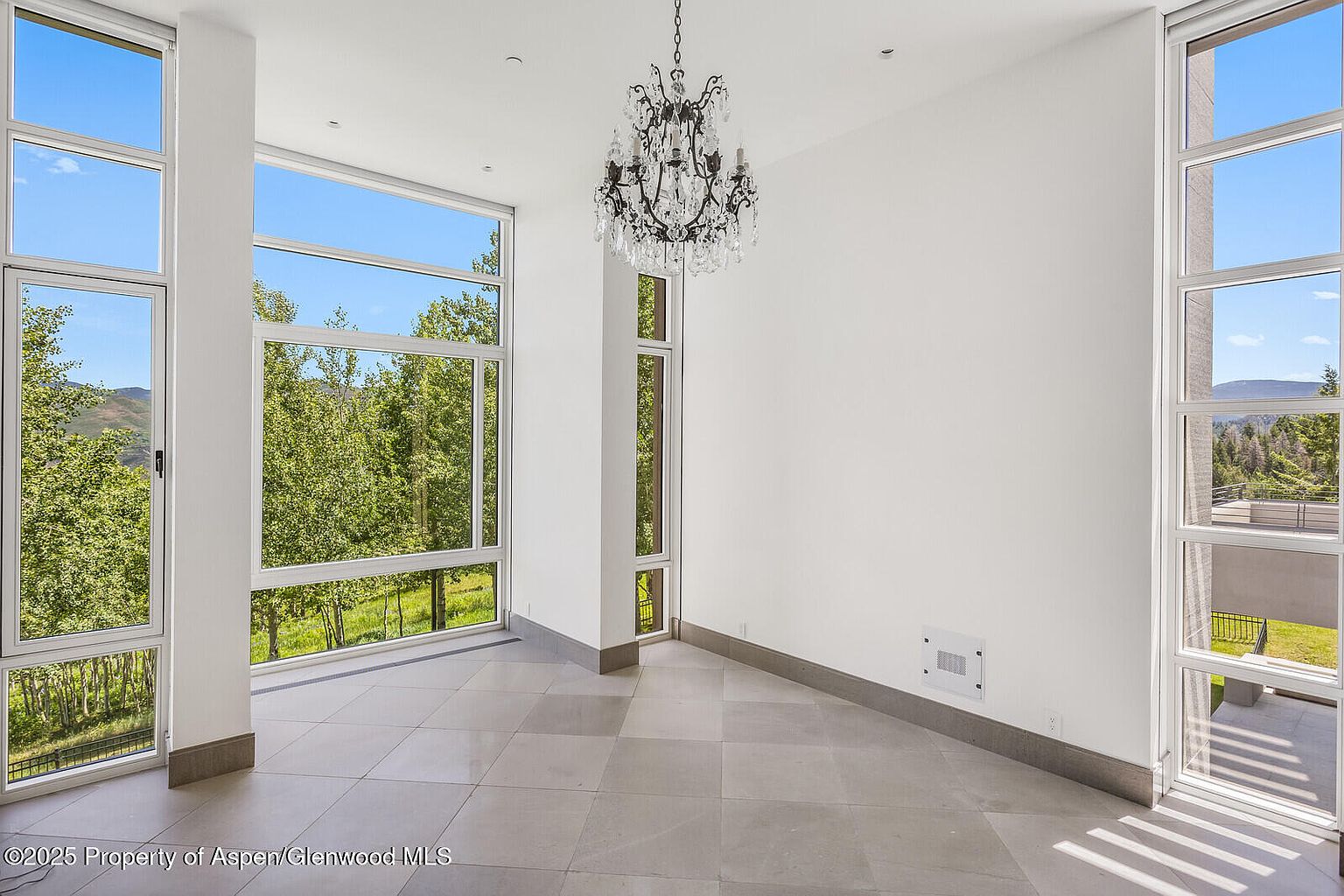 This is an interior shot of a modern living room featuring large windows that offer views of lush greenery. A decorative chandelier hangs from the ceiling, adding a touch of elegance to the minimalist space. The room has light-colored tile flooring and white walls, creating a bright and airy atmosphere.