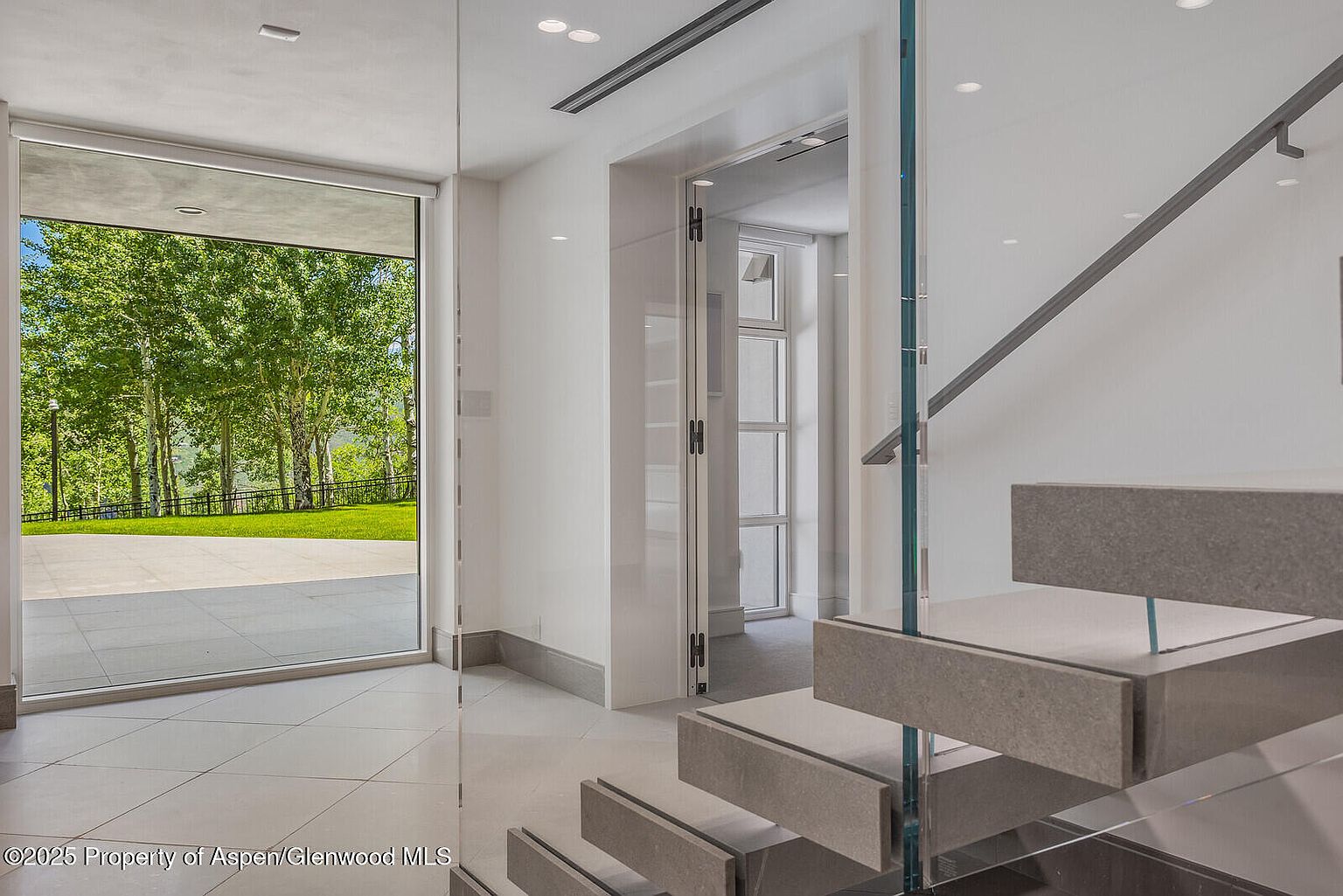 This interior shot showcases a modern hallway with floating stairs, featuring stone treads and glass railings. A large glass door provides a view to the exterior, while another doorway leads to a different room. The space is bright and airy, with clean lines and a minimalist aesthetic, creating a sophisticated and inviting atmosphere.