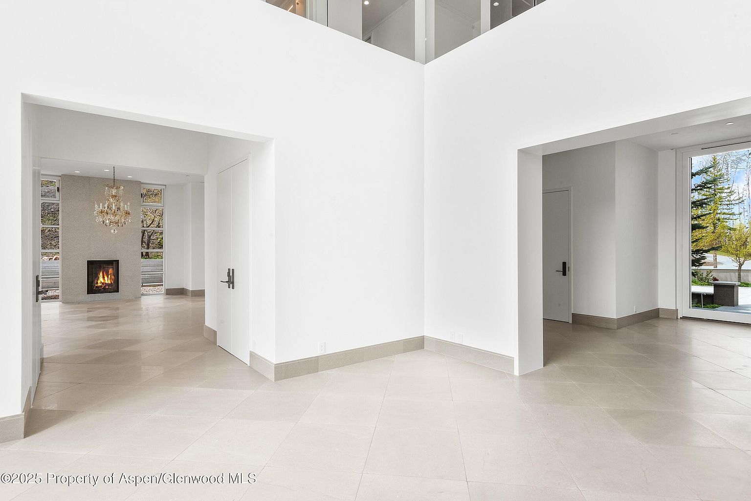 This interior shot showcases a spacious hallway with a modern aesthetic, featuring light-colored tile flooring and crisp white walls. The hallway branches into different areas, with doorways leading to other rooms and a glimpse of a fireplace in one direction and an outdoor view through a large window in another. The high ceilings and minimalist design contribute to an open and airy feel, emphasizing the home's contemporary style.