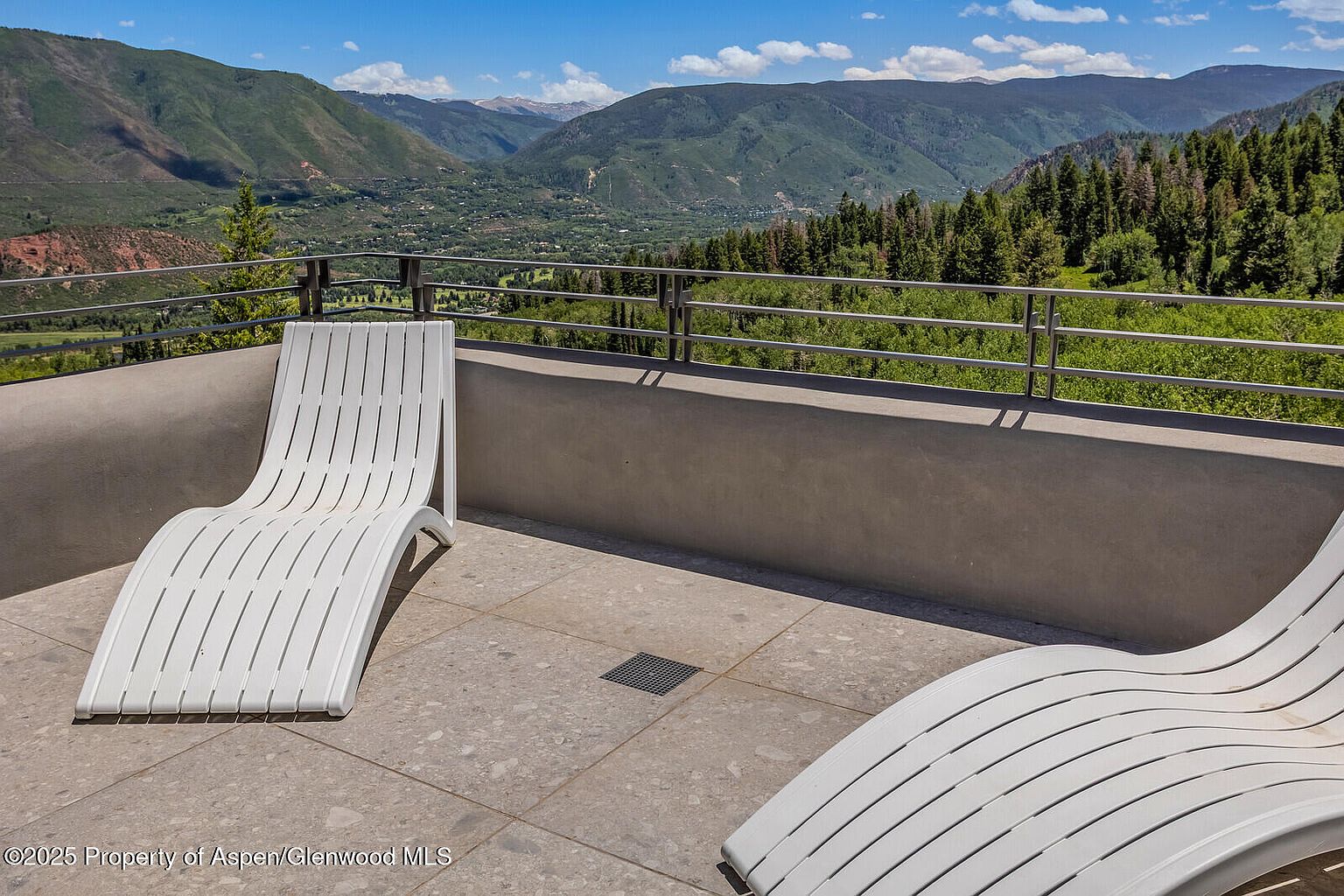 This image showcases a modern patio or balcony with stunning mountain views. Two sleek, white lounge chairs are positioned on a tiled surface, suggesting a relaxing outdoor space. A minimalist metal railing and a low concrete wall provide safety and definition to the area, while the backdrop features lush green mountains under a clear blue sky, enhancing the property's appeal.