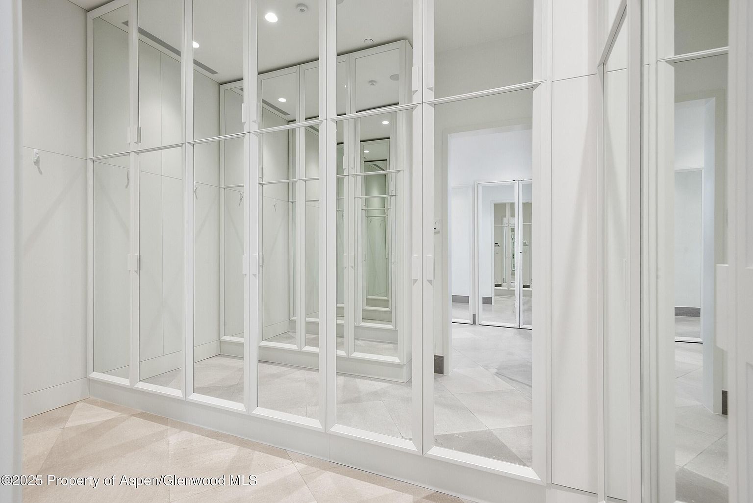 This is a bright and spacious walk-in closet featuring custom white cabinetry and mirrored doors. The closet includes ample storage space with shelving and hanging rods. The flooring is a light-colored tile, and the overall design is clean and modern, creating a luxurious and organized space.