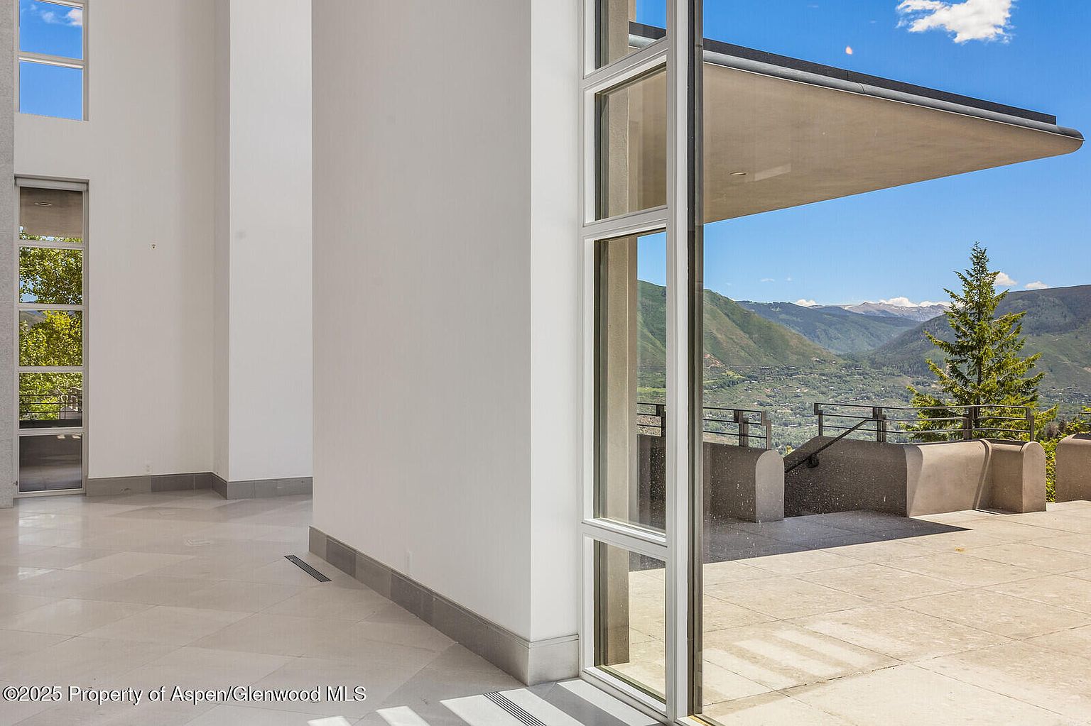 The image showcases a modern hallway with large windows offering a stunning view of the mountains. The interior features clean lines, white walls, and light-colored tile flooring, creating a bright and airy atmosphere. The open doorway leads to an outdoor patio or deck area, seamlessly blending indoor and outdoor living spaces.