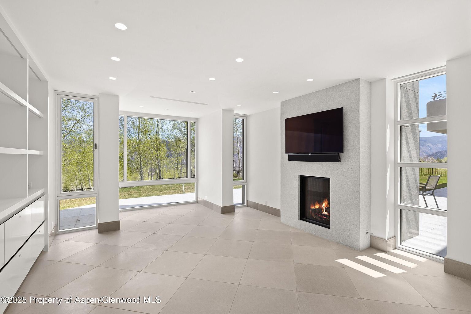 This is a bright and spacious living room featuring large windows that offer scenic views of the outdoors. The room is minimally furnished, highlighting the tile flooring and a modern fireplace with a mounted television. Built-in shelving adds to the room's functionality and clean aesthetic.