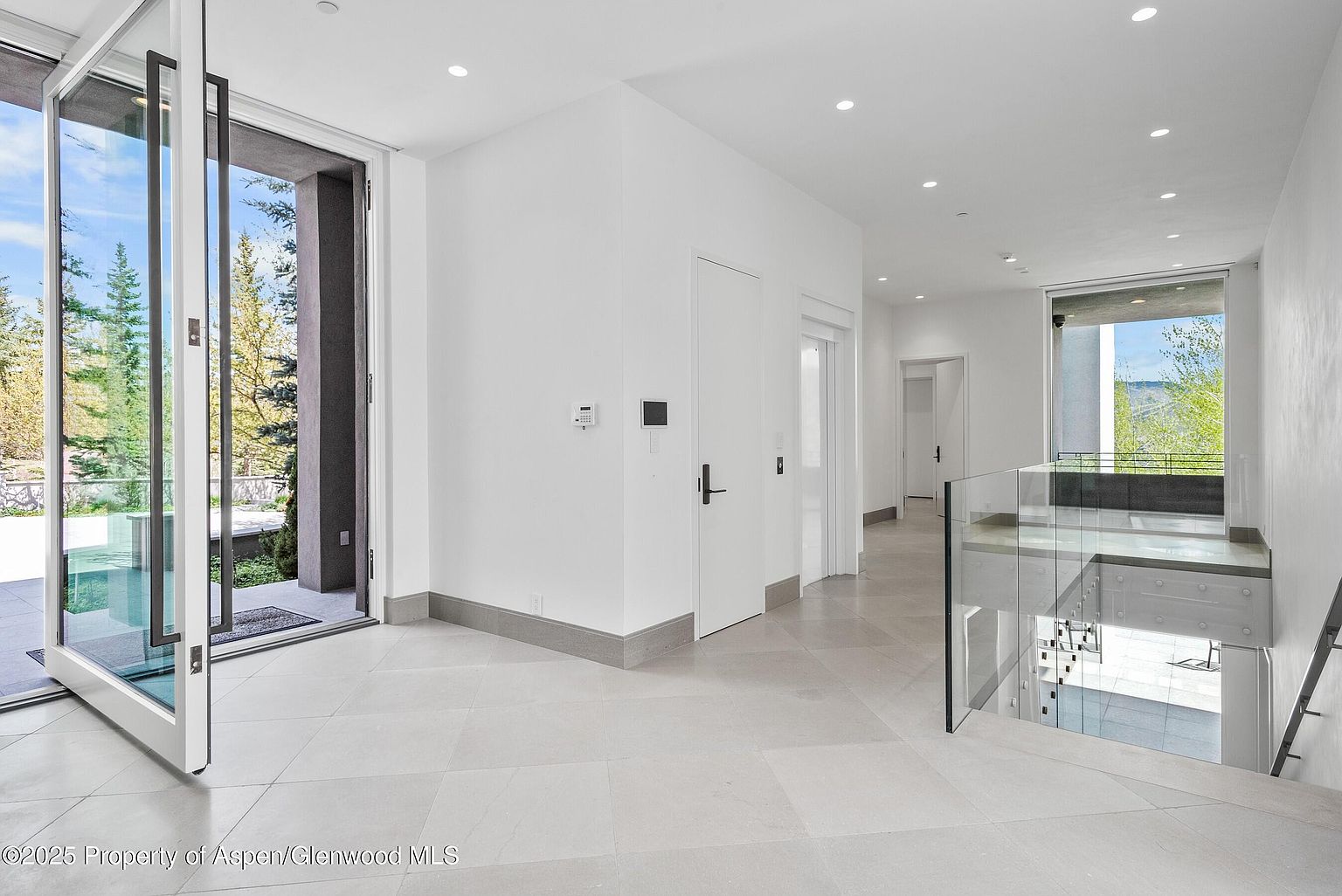 This interior shot showcases a bright and modern hallway with a staircase. The hallway features light tile flooring, white walls, and recessed lighting, creating a clean and spacious feel. A glass railing provides a contemporary touch to the staircase, while large windows and doors offer views of the outdoors, enhancing the natural light and connection to the surrounding landscape.