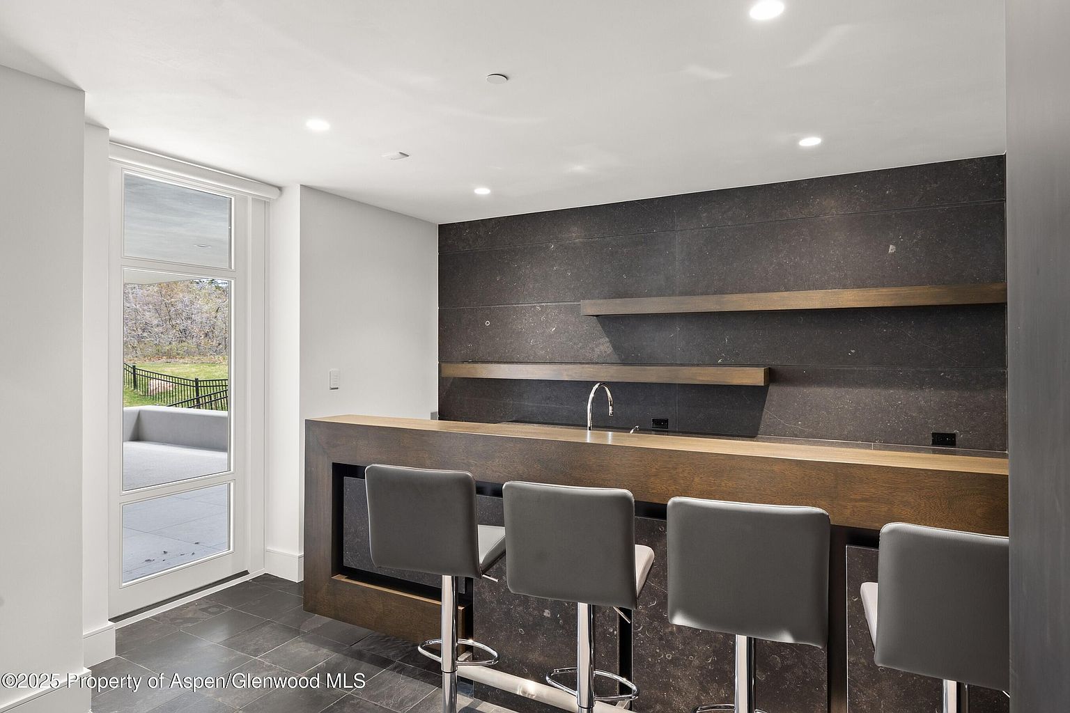 This interior shot showcases a modern bar area with a dark wood countertop and sleek gray bar stools. The backsplash is a textured dark stone, complemented by floating wooden shelves. A door to the outside provides natural light and a view of the outdoor space.