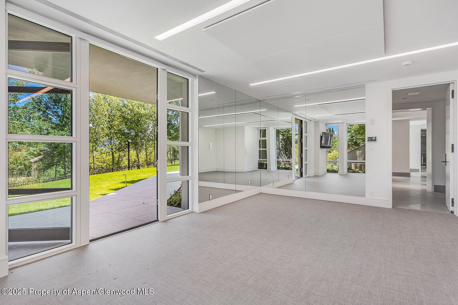 This is a spacious home gym featuring mirrored walls, large windows offering views of the outdoors, and light-colored flooring. The room is bright and airy, with modern lighting fixtures and an open layout that provides ample space for exercise equipment and activities. The overall impression is clean, contemporary, and conducive to a healthy lifestyle.