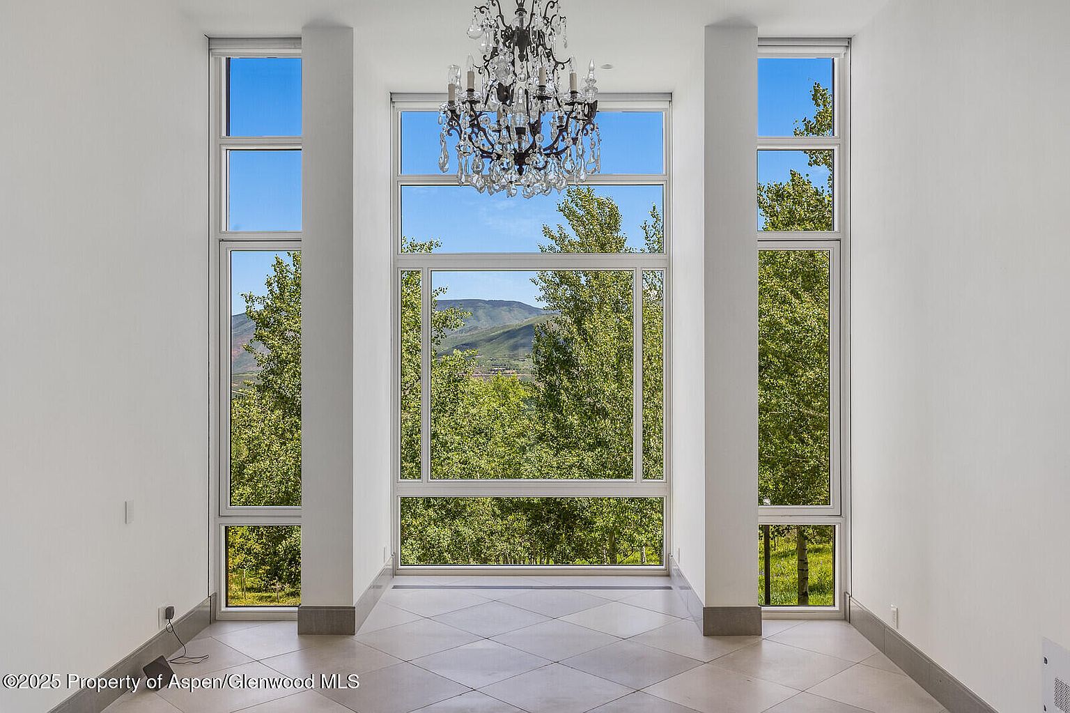 This is an interior shot of a living room featuring a wall of large windows offering a view of lush greenery and a distant mountain. A decorative chandelier hangs from the ceiling, adding a touch of elegance to the space. The room is spacious with white walls and tiled floors, creating a bright and airy atmosphere.