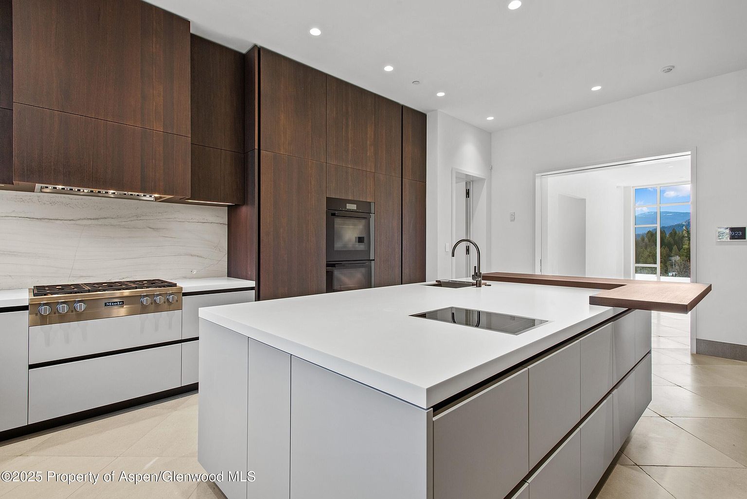This is a modern kitchen featuring dark wood cabinetry and a large white island with a built-in cooktop. The kitchen is well-lit with recessed lighting, and a window in the background offers a view of the outdoors. The overall impression is sleek and contemporary.