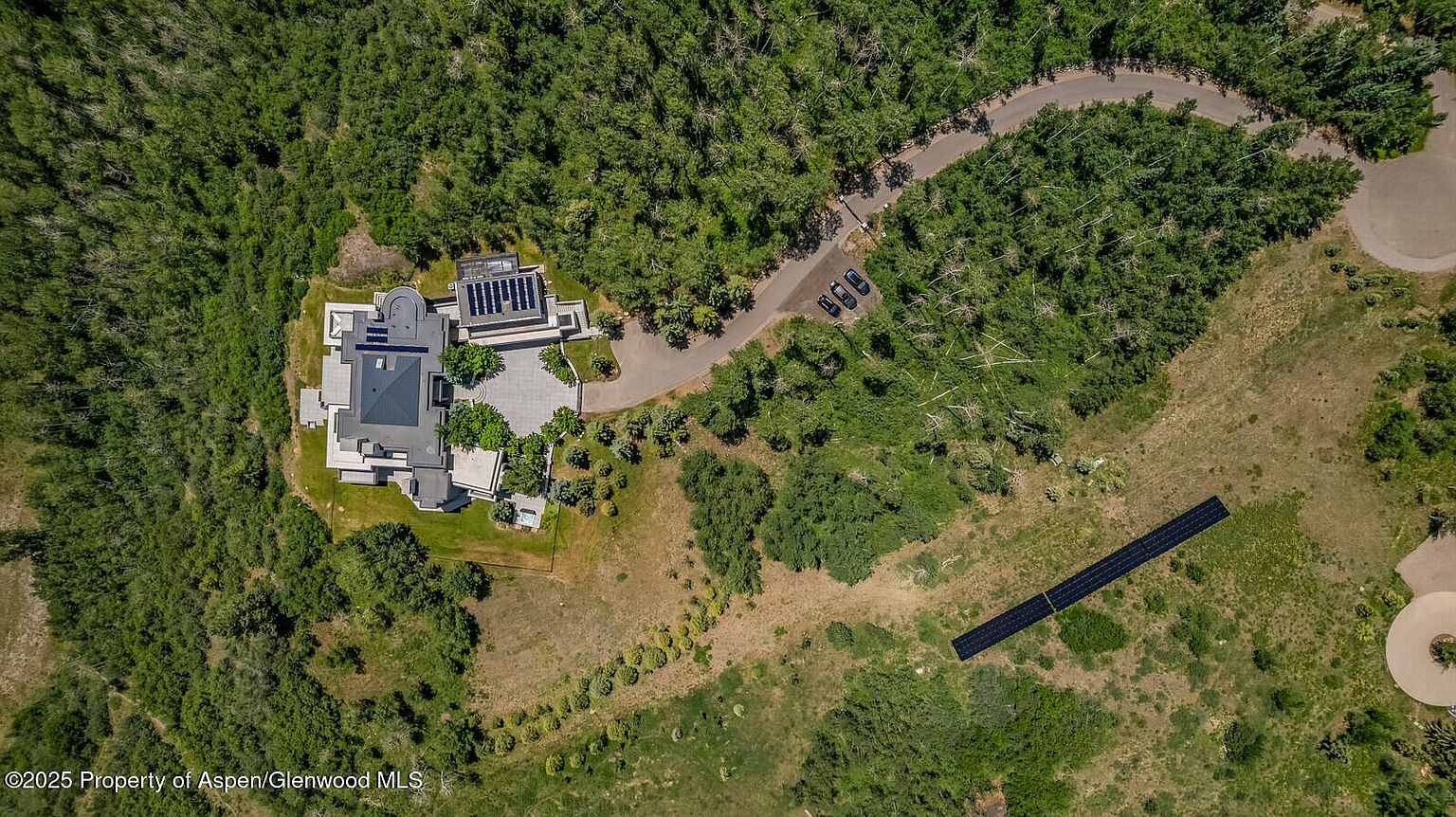 This aerial view showcases a luxurious estate nestled amidst lush greenery. The property features a modern architectural design with a dark roof, solar panels, and meticulously landscaped grounds. A winding driveway leads to the house, with several cars parked near the entrance, and a separate solar panel array is visible in the yard.