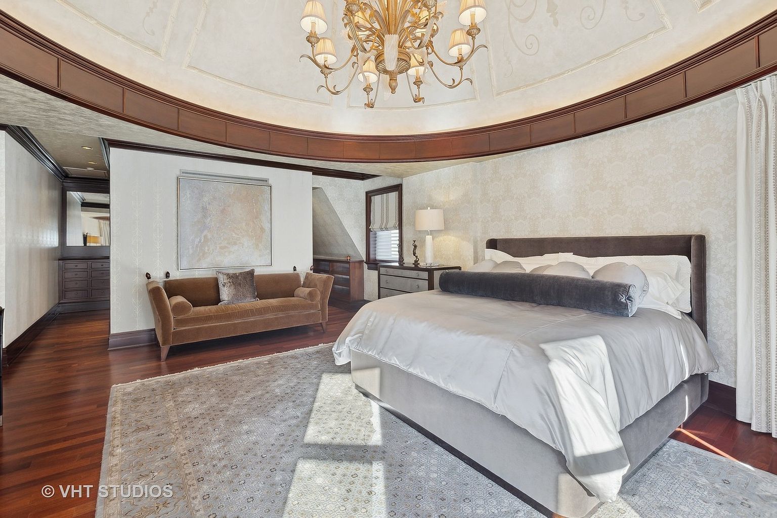 This is a luxurious primary bedroom featuring a large bed with a gray headboard and white linens, complemented by a gray bolster pillow. The room has a unique architectural design with a domed ceiling and a decorative chandelier. A brown sofa sits against the wall, and a patterned rug covers the hardwood floor, adding warmth and texture to the space.