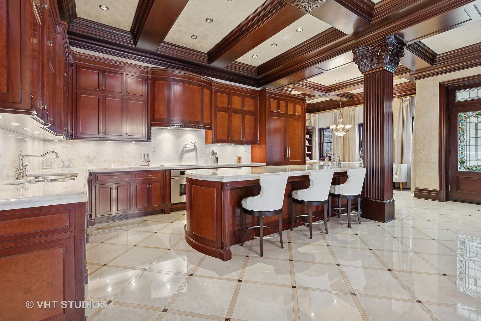 This is a luxurious kitchen featuring dark wood cabinetry, marble countertops, and a large island with seating. The coffered ceiling with dark wood beams adds a touch of elegance, while the tiled floor complements the overall design. A decorative column stands near the island, adding to the grandeur of the space.