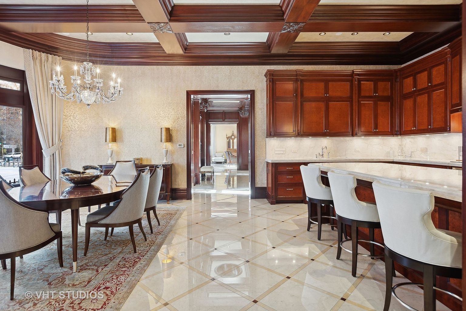 This interior shot showcases an elegant dining area with a dark wood table surrounded by upholstered chairs, illuminated by a crystal chandelier. The room features a patterned wallpaper, rich wood trim, and a marble tile floor, creating a luxurious and sophisticated atmosphere. A doorway leads to another room, adding depth and visual interest.