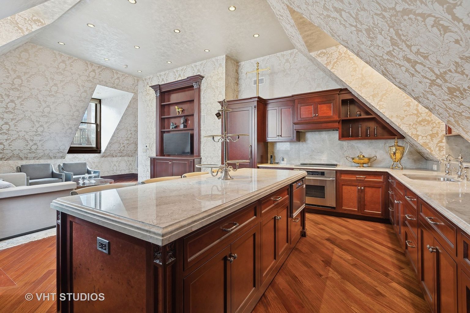 This is an interior shot of a luxurious kitchen featuring dark wood cabinetry, marble countertops, and hardwood floors laid in a herringbone pattern. The kitchen island is the focal point, complemented by built-in shelving and modern appliances. The room is well-lit with recessed lighting, and the wallpaper adds a touch of elegance.