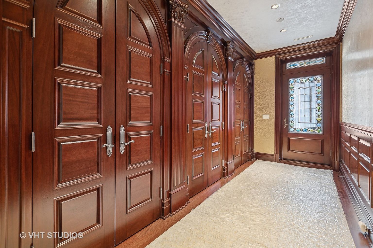 This interior shot showcases a grand hallway featuring rich, dark wood doors with arched tops and ornate detailing. The walls are partially paneled, and a decorative stained-glass door is visible at the end of the hall. A light-colored rug runs the length of the hallway, adding a touch of elegance.