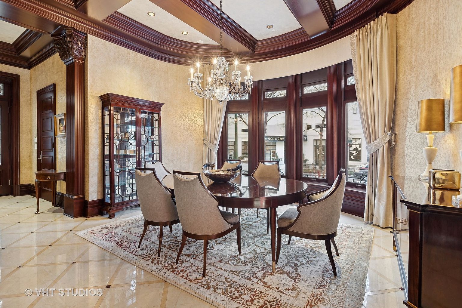 This is an elegant dining room featuring a round dark wood table surrounded by six upholstered chairs. A crystal chandelier hangs above the table, and a large window with draped curtains provides natural light. The room is decorated with a patterned rug and a display cabinet, creating a sophisticated and inviting atmosphere.