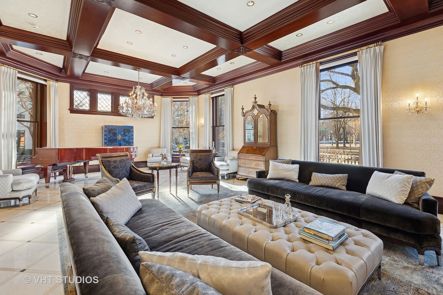This is a luxurious living room featuring a coffered ceiling with dark wood beams, a large crystal chandelier, and elegant furnishings. The room includes two sofas, a tufted ottoman, and several armchairs, creating a comfortable and sophisticated space. Large windows with sheer curtains provide ample natural light, highlighting the room's refined details and classic style.
