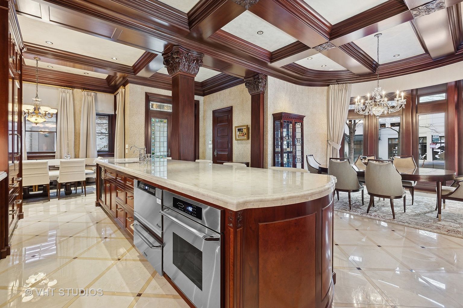 This is a luxurious kitchen featuring a large island with a marble countertop and stainless steel appliances. The room is adorned with dark wood cabinetry and columns, complemented by a coffered ceiling with intricate detailing. The flooring is polished tile, and the space opens to a dining area with a chandelier, creating an elegant and inviting atmosphere.