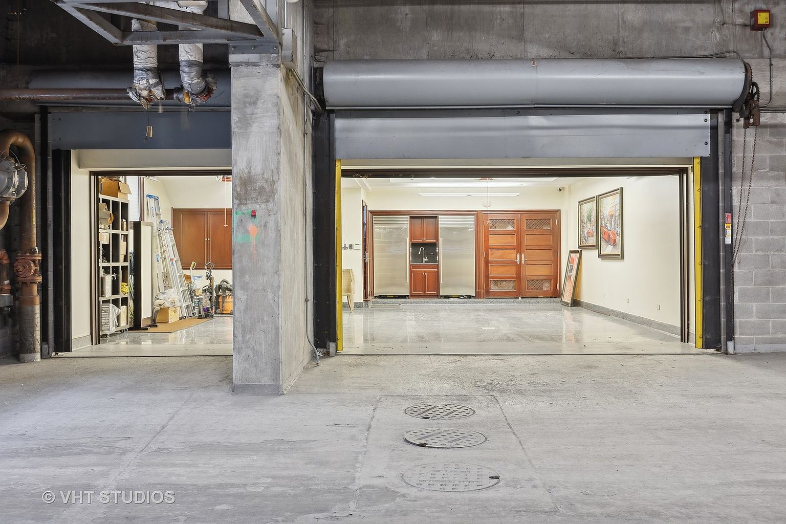 This is an interior shot of a garage space, featuring two large roll-up doors that lead to separate interior rooms. The garage has a concrete floor with visible utility access points. The interior rooms visible through the garage doors appear to be finished spaces, one possibly a storage area and the other a kitchen or dining area.