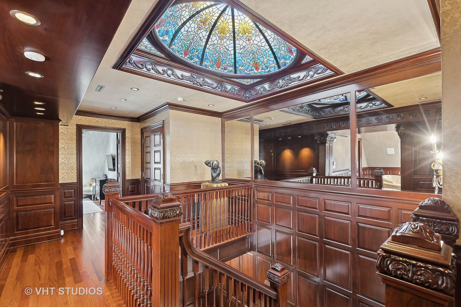 This interior shot showcases a luxurious hallway and staircase area, featuring rich wood paneling and a decorative stained-glass skylight. The space is illuminated by recessed lighting and natural light filtering through the skylight, creating a warm and inviting atmosphere. A mirrored wall reflects the staircase and adds depth to the space, enhancing the overall elegance and grandeur.
