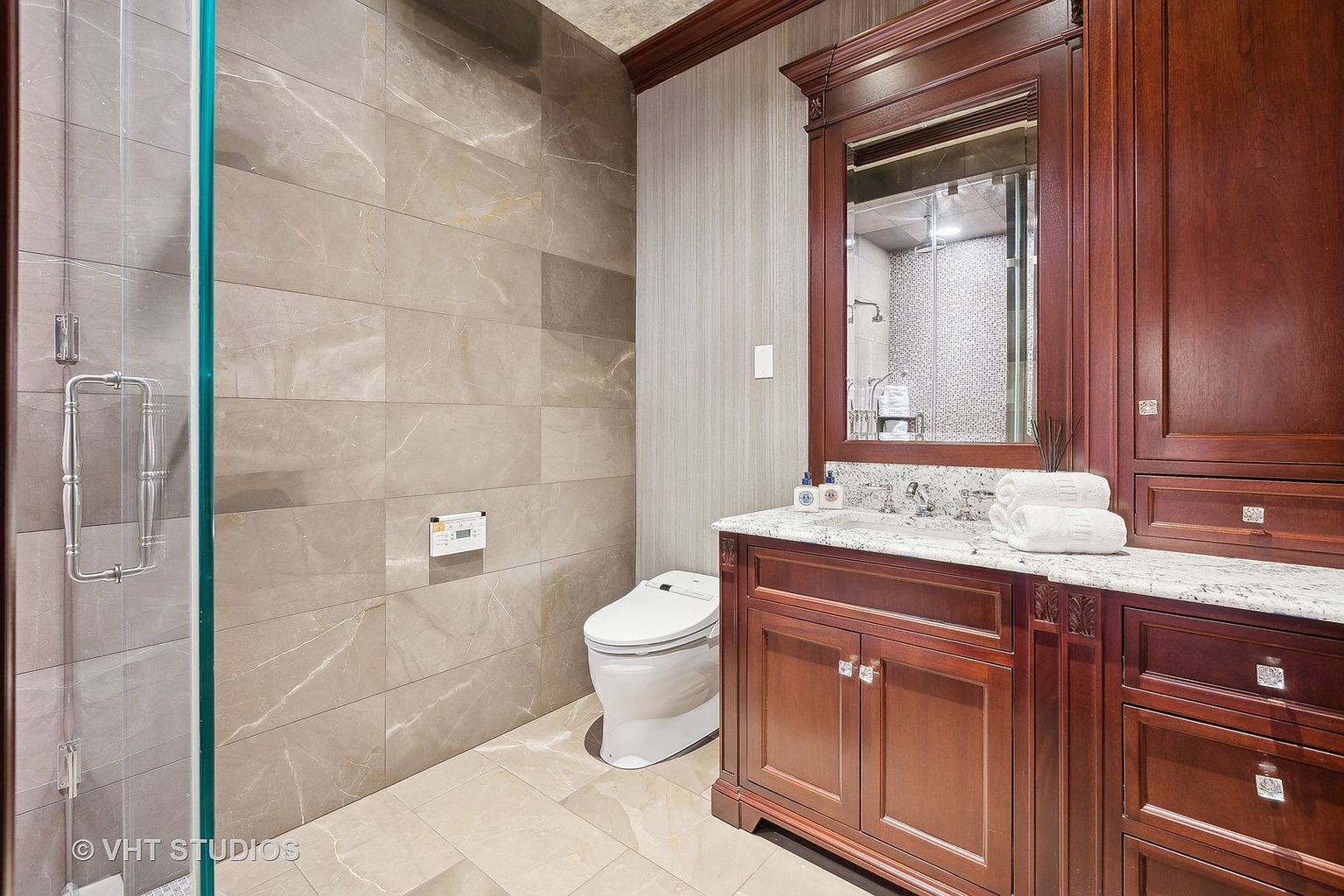 This is a primary bathroom featuring a glass-enclosed shower, a modern toilet, and a vanity with a dark wood cabinet and a light-colored countertop. The walls are tiled with large, neutral-toned tiles, and the overall style is luxurious and contemporary. The perspective is from the doorway, showcasing the entire bathroom.