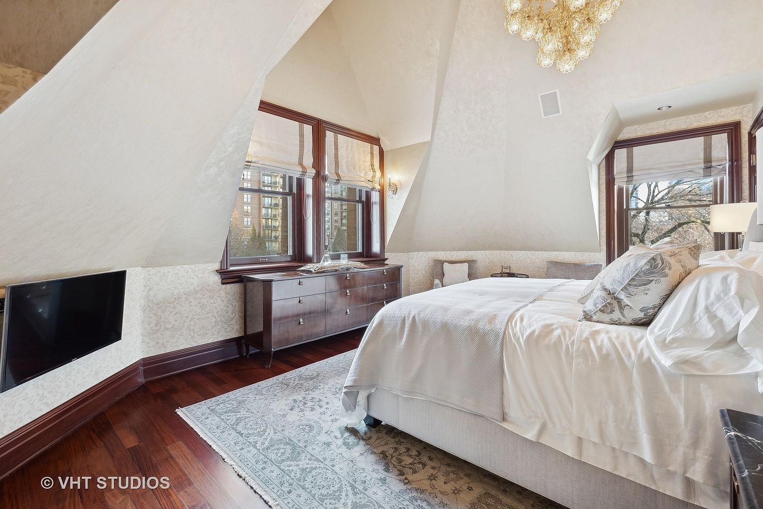 This is a primary bedroom featuring a large bed with white linens and decorative pillows, a dresser under a window with roman shades, and a wall-mounted television. The room has an attic-style ceiling and a patterned rug on hardwood floors. The overall impression is luxurious and comfortable.
