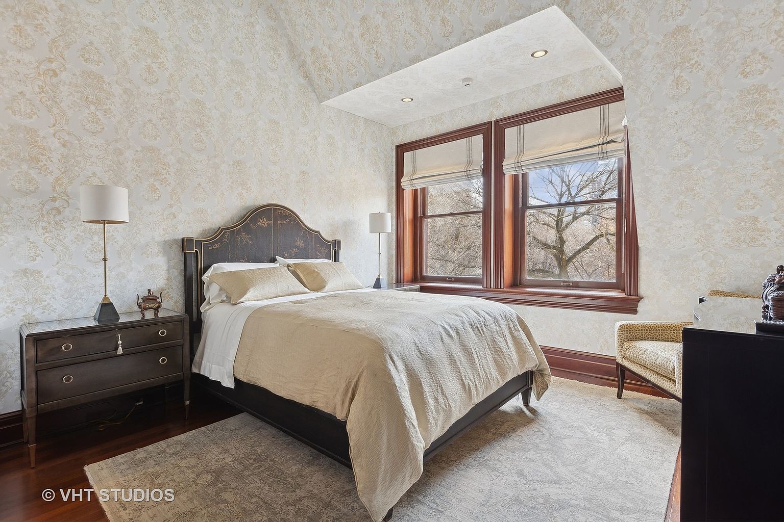 This is a primary bedroom featuring a bed with a decorative headboard, a nightstand with a lamp, and a window with blinds. The walls are covered in patterned wallpaper, and the room is furnished with a chair and a dresser. The perspective is from the doorway, showcasing the room's layout and design.