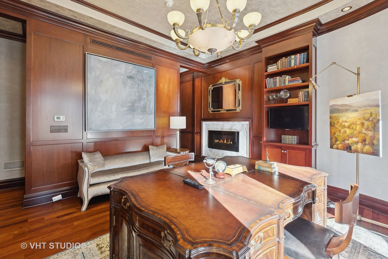 This is a luxurious home office featuring rich wood paneling, a large ornate desk, and a cozy seating area with a fireplace. The room is well-lit by a chandelier and a table lamp, creating a warm and inviting atmosphere. A built-in bookcase and artwork add to the sophisticated ambiance, making it an ideal space for work and relaxation.